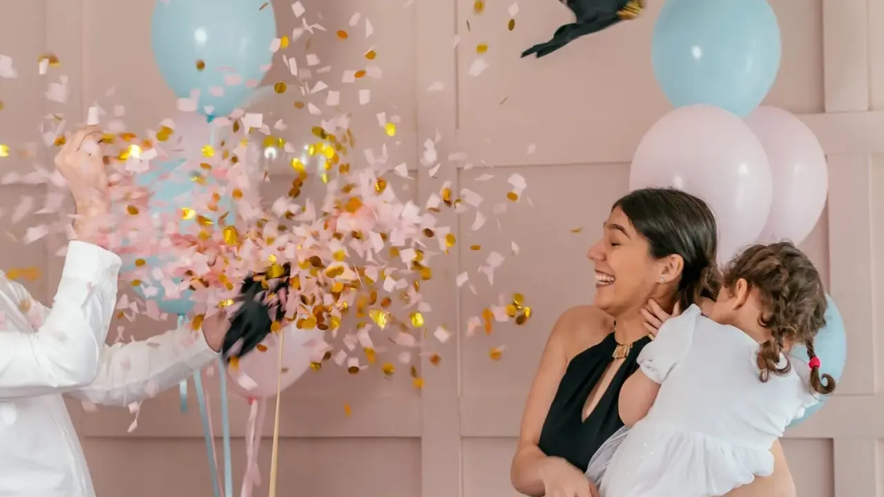 Explosion de confettis roses et dorés lors d'une fête pour annoncer fille ou garçon. Une femme rit, tenant un enfant, entourée de ballons bleus et roses.