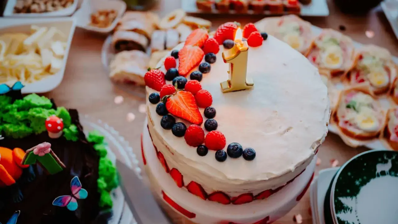 Gâteau à deux étages décoré de fruits rouges et d'une bougie "1" pour un déjeuner d'anniversaire. Un autre gâteau chocolaté, des petits fours et des fruits sont disposés sur la table.