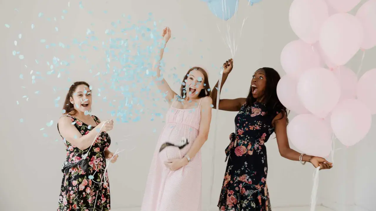 Trois femmes célèbrent un gender reveal, avec des confettis bleus et des ballons roses et bleus. C'est quoi ce moment joyeux !