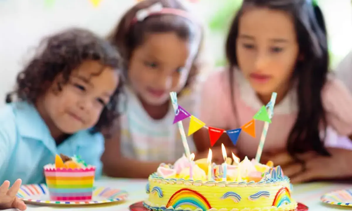 Trois enfants regardent un gâteau d'anniversaire jaune décoré d'arcs-en-ciel et de fanions. Le meilleur goût gâteau anniversaire est prêt à être dégusté.