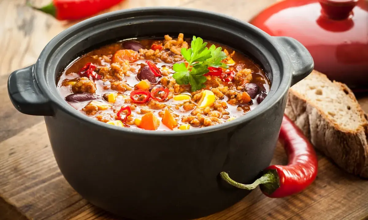 Un plat de chili mijoté dans une cocotte inox four, garni de persil, piments et maïs. Parfait pour un repas réconfortant.