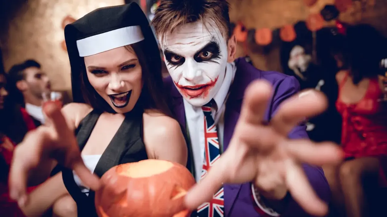 Un couple en costumes effrayants, une nonne et le Joker, profite d'une **soirée costumée** d'Halloween, tenant une citrouille sculptée.