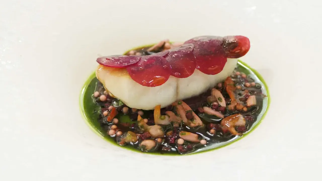 Un poisson blanc délicat, garni de fines lamelles de betterave rouge, repose sur un lit de grains de riz noir et de morceaux de poulpe, le tout nappé d'une sauce verte vibrante. Une œuvre d'art culinaire, un de ces mets d'exception.