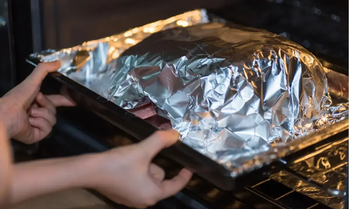 Mains dans le four, une personne place un plat recouvert de papier **alu** pour une cuisson parfaite.