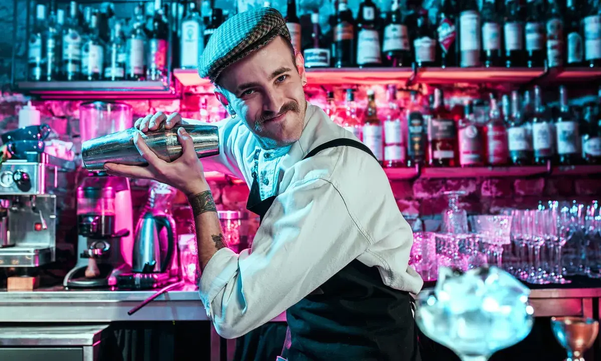 Un barman souriant, casquette vissée, montre comment utiliser un shaker. Derrière lui, des étagères remplies de bouteilles s'illuminent de néons roses et bleus.