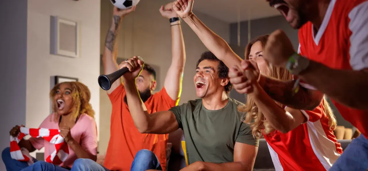 Des supporters en pleine soiree foot, exultant devant leur écran. Un homme lève un ballon, un autre souffle dans une trompette.