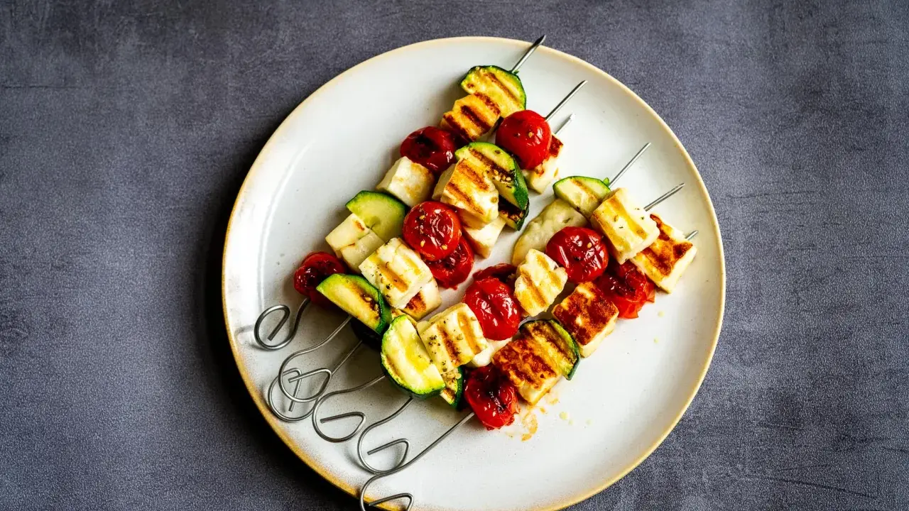 Brochettes de barbecue végétarien : cubes de halloumi grillé, tomates cerises et courgettes sur des brochettes métalliques, servis sur une assiette.
