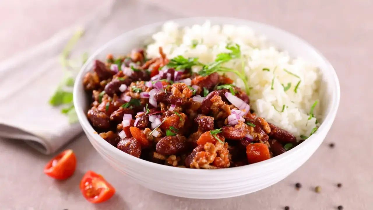 Un bol de riz blanc et de chili con carne recette de chef, garni d'oignons rouges et de persil. Des tomates cerises et du poivre en grains décorent la table.