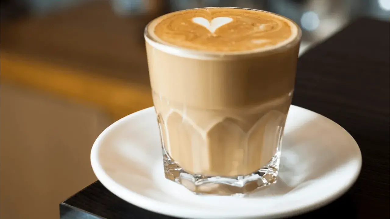 Un café latte crémeux avec un cœur dessiné dans la mousse, servi dans un verre transparent sur une soucoupe blanche.