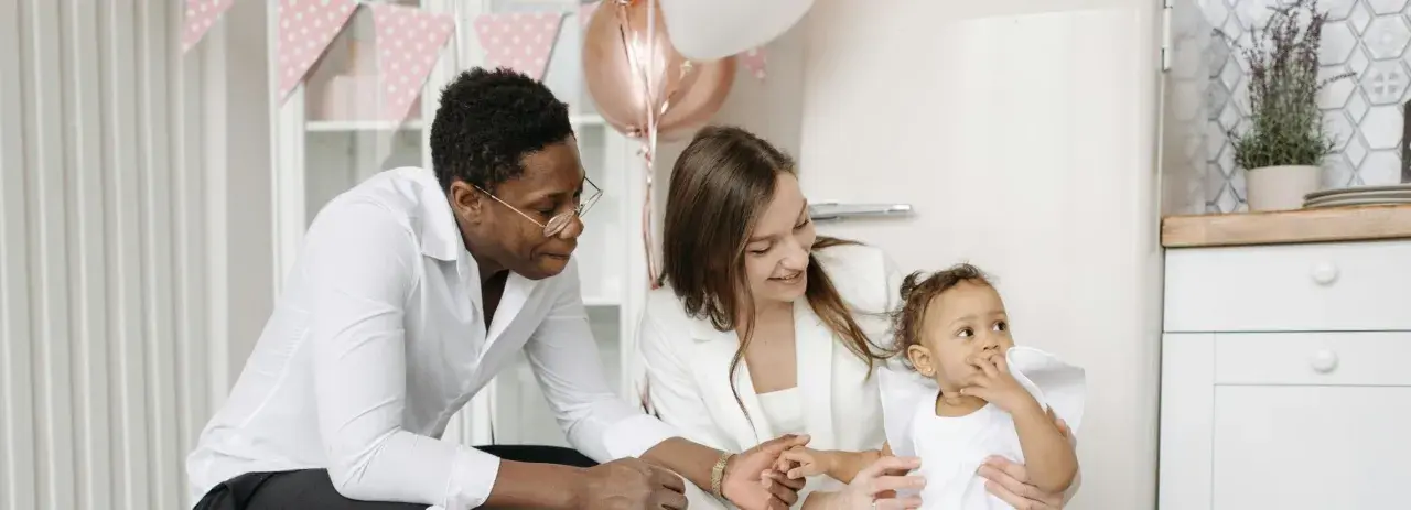 Famille souriante préparant une fête. La mère tient le bébé, le père regarde. Décoration de ballons et guirlandes roses. Une aide pour le check list bapteme.