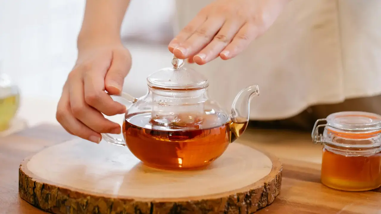 Mains tenant une théière en verre remplie d'un thé ambré, prête à servir le meilleur thé. Un pot de miel à côté.