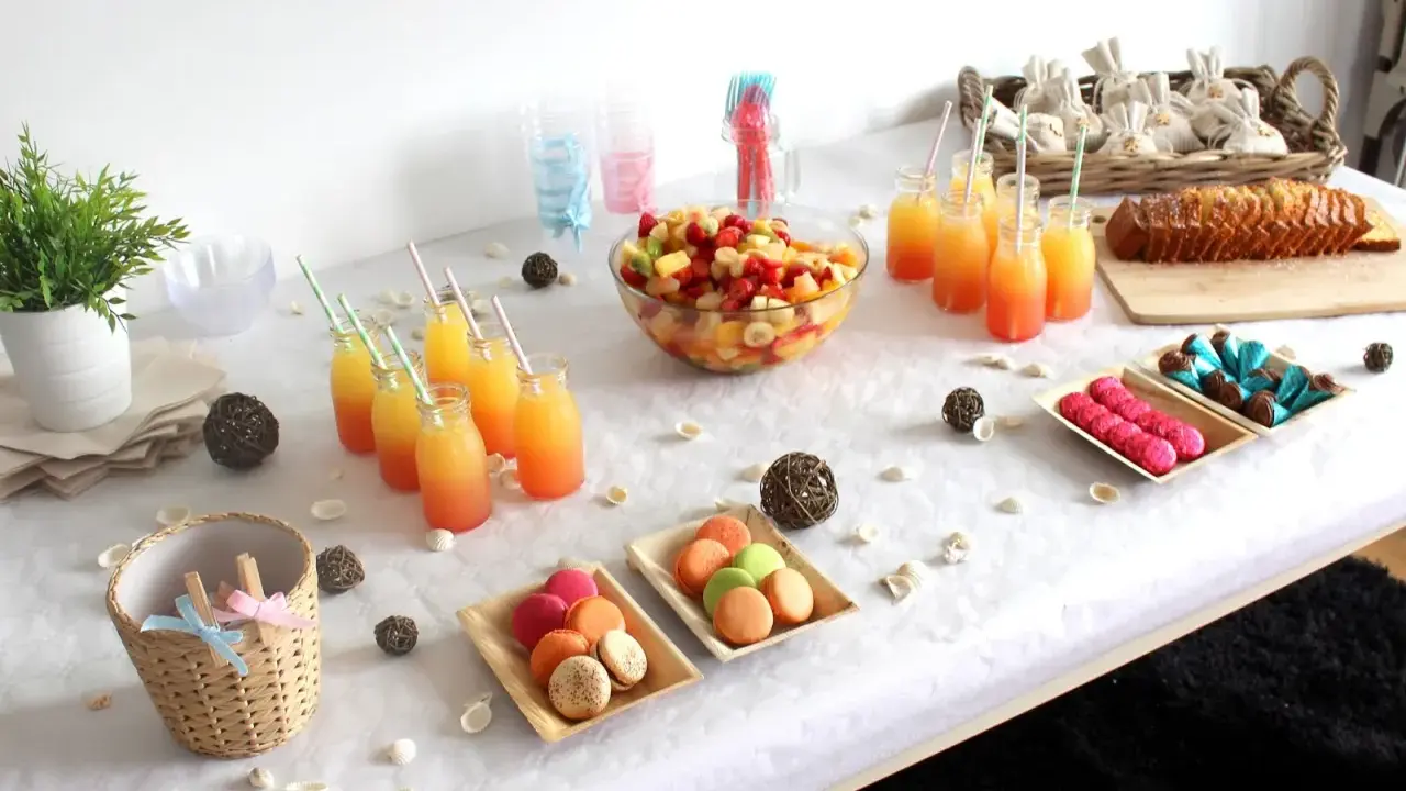 Buffet d'une idée gender reveal : jus de fruits colorés, salade de fruits, macarons roses et verts, gâteau, chocolats bleus et roses.