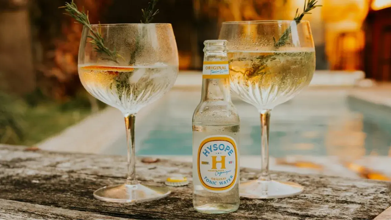 Une bouteille de tonic Hysopé Original, prête à être versée dans deux verres de gin rafraîchissants, décorés de romarin et d'une tranche d'orange.