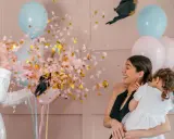 Explosion de confettis roses et dorés lors d'une fête pour annoncer fille ou garçon. Une femme rit, tenant un enfant, entourée de ballons bleus et roses.