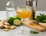Un cocktail gingembre rafraîchissant, garni d'une tranche de citron vert, avec du gingembre frais, des citrons verts et de la menthe disposés autour.