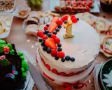 Gâteau à deux étages décoré de fruits rouges et d'une bougie "1" pour un déjeuner d'anniversaire. Un autre gâteau chocolaté, des petits fours et des fruits sont disposés sur la table.