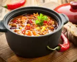 Un plat de chili mijoté dans une cocotte inox four, garni de persil, piments et maïs. Parfait pour un repas réconfortant.