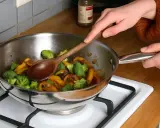Une personne remue des légumes dans une poêle en inox alimentaire avec une cuillère en bois.
