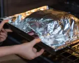 Mains dans le four, une personne place un plat recouvert de papier **alu** pour une cuisson parfaite.