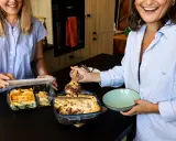 Deux femmes souriantes servent un plat gourmand d'une casserole qui va au four. L'une sert une lasagne, l'autre un dessert chocolaté.