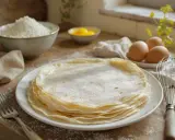Une pile de crêpes de riz fines et dorées, saupoudrées de sucre glace, prêtes à être dégustées. Des ingrédients pour les préparer sont disposés autour.