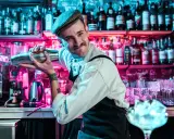 Un barman souriant, casquette vissée, montre comment utiliser un shaker. Derrière lui, des étagères remplies de bouteilles s'illuminent de néons roses et bleus.