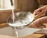 Main d'une personne nettoyant une poêle Tefal avec du vinaigre blanc et une éponge sous un robinet d'eau savonneuse.
