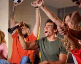 Des supporters en pleine soiree foot, exultant devant leur écran. Un homme lève un ballon, un autre souffle dans une trompette.