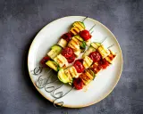 Brochettes de barbecue végétarien : cubes de halloumi grillé, tomates cerises et courgettes sur des brochettes métalliques, servis sur une assiette.