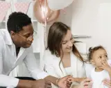 Famille souriante préparant une fête. La mère tient le bébé, le père regarde. Décoration de ballons et guirlandes roses. Une aide pour le check list bapteme.