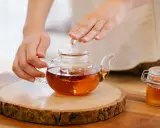 Mains tenant une théière en verre remplie d'un thé ambré, prête à servir le meilleur thé. Un pot de miel à côté.