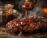 Un pinceau dépose une sauce pour barbecue brillante et fumante sur un plat de viande grillée. Un pot de sauce est visible en arrière-plan.