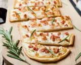 Quatre tartes flambées, une délicieuse entrée alsacienne, garnies de lardons et de romarin sur une planche en bois.