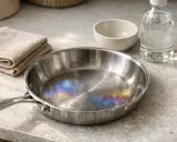 Casserole inox avec des taches bleues iridescentes sur le fond, à côté d'une serviette pliée et d'une bouteille de liquide.