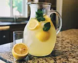 Un pichet de citronnade maison rafraîchissante, garni de citrons et de menthe, prêt à être servi avec un verre et un demi-citron sur un comptoir.