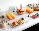 Buffet d'une idée gender reveal : jus de fruits colorés, salade de fruits, macarons roses et verts, gâteau, chocolats bleus et roses.