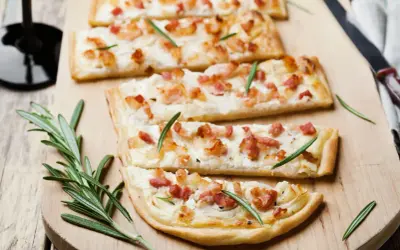Quatre tartes flambées, une délicieuse entrée alsacienne, garnies de lardons et de romarin sur une planche en bois.