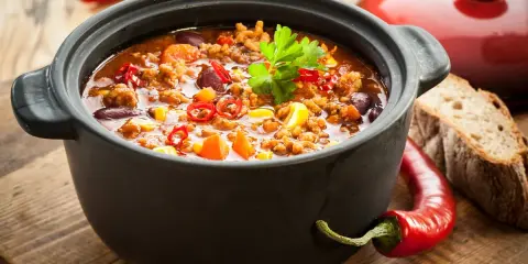 Un plat de chili mijoté dans une cocotte inox four, garni de persil, piments et maïs. Parfait pour un repas réconfortant.