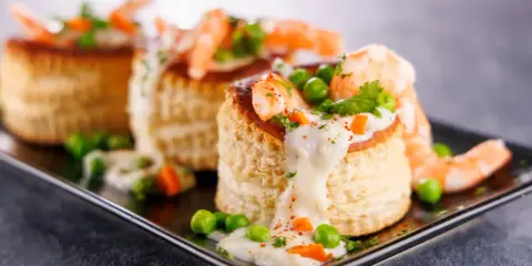 Trois bouchées de tapas crevette, garnies de crevettes, petits pois et sauce crémeuse, sur un plateau noir.