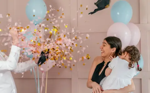 Explosion de confettis roses et dorés lors d'une fête pour annoncer fille ou garçon. Une femme rit, tenant un enfant, entourée de ballons bleus et roses.