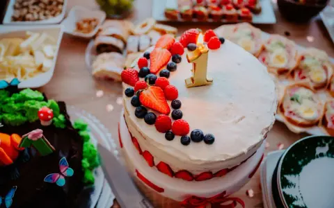 Gâteau à deux étages décoré de fruits rouges et d'une bougie "1" pour un déjeuner d'anniversaire. Un autre gâteau chocolaté, des petits fours et des fruits sont disposés sur la table.
