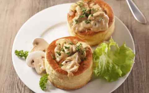 Deux bouchées à la reine maison, garnies d'une sauce crémeuse aux champignons et persil, servies avec des champignons frais et une feuille de laitue.