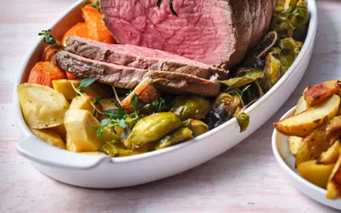 Tranches de rosbeef froid, accompagnées de pommes de terre rôties, carottes et choux de Bruxelles dans un plat blanc.