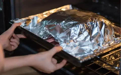 Mains dans le four, une personne place un plat recouvert de papier **alu** pour une cuisson parfaite.