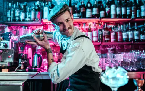 Un barman souriant, casquette vissée, montre comment utiliser un shaker. Derrière lui, des étagères remplies de bouteilles s'illuminent de néons roses et bleus.