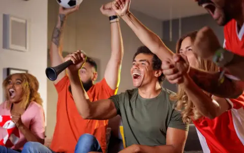 Des supporters en pleine soiree foot, exultant devant leur écran. Un homme lève un ballon, un autre souffle dans une trompette.
