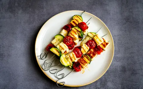 Brochettes de barbecue végétarien : cubes de halloumi grillé, tomates cerises et courgettes sur des brochettes métalliques, servis sur une assiette.