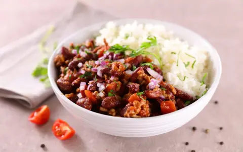 Un bol de riz blanc et de chili con carne recette de chef, garni d'oignons rouges et de persil. Des tomates cerises et du poivre en grains décorent la table.