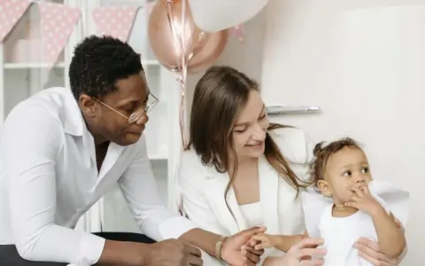 Famille souriante préparant une fête. La mère tient le bébé, le père regarde. Décoration de ballons et guirlandes roses. Une aide pour le check list bapteme.