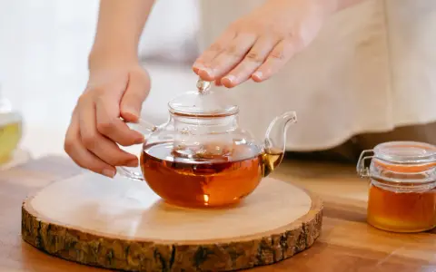 Mains tenant une théière en verre remplie d'un thé ambré, prête à servir le meilleur thé. Un pot de miel à côté.